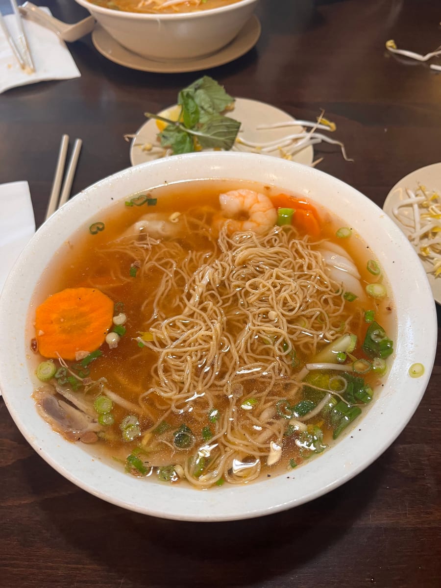 A bowl of egg noodle soup with shrimp, carrot and green onion on a dark table